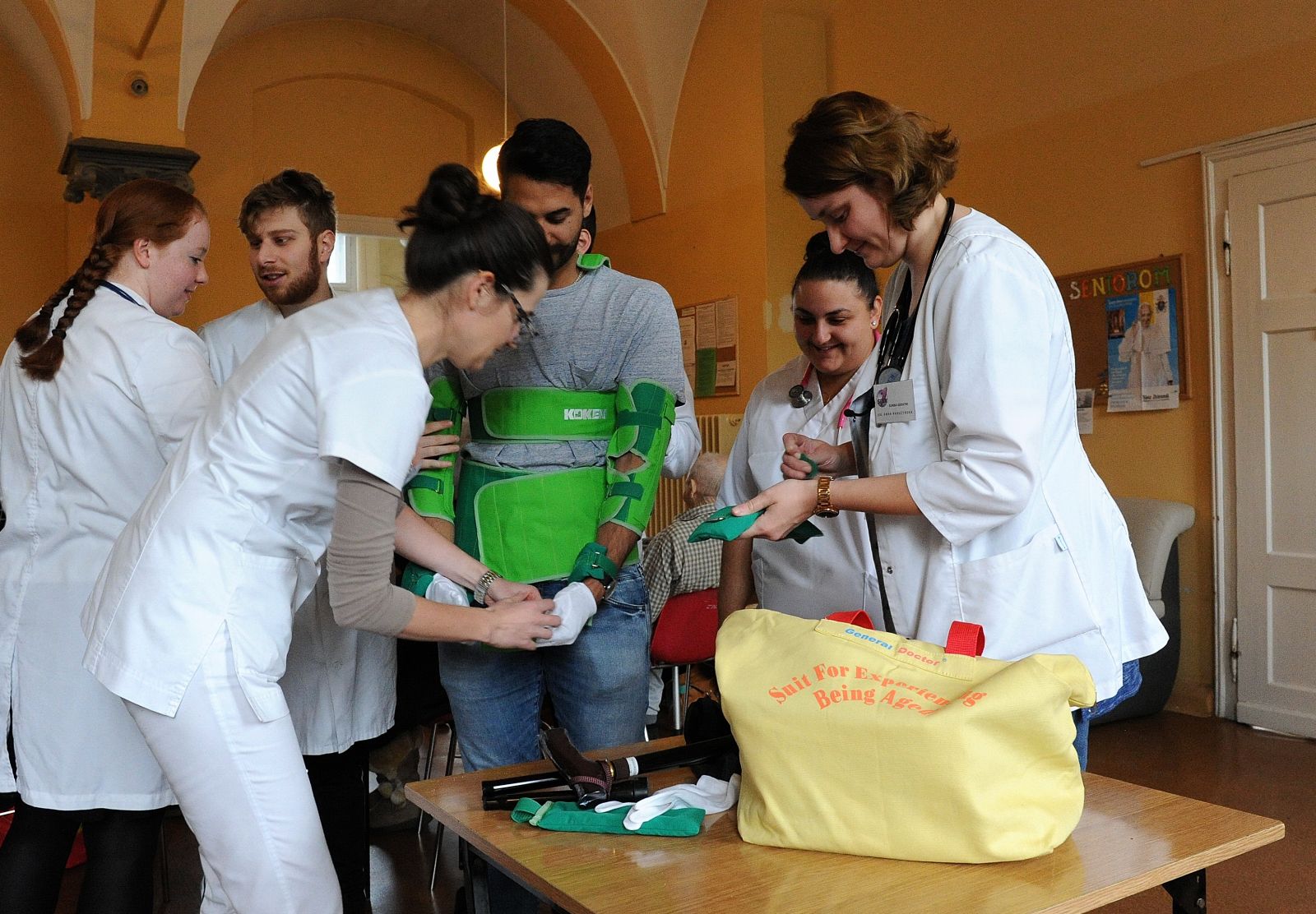 Zdjęcie w sali szpitalnej. Dwie lekarki ubierają studenta w strój medyczny. Dwie studentki się przyglądają.