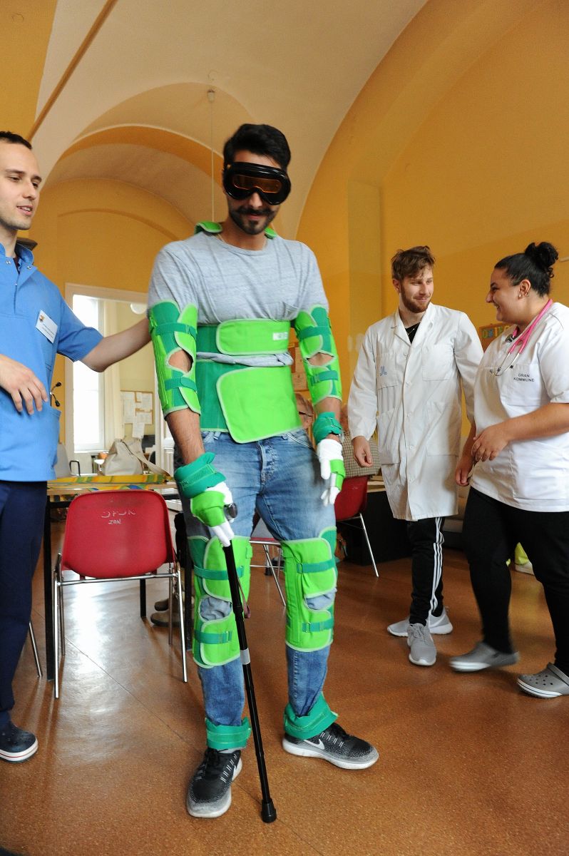 Zdjecie zrobione w sali szpitalnej. Na środku zdjęcia stoi student ubrany w strój medyczny. Po lewej stronie studenta ubranego w strój medyczny stoi lekarz i podpiera go ręką. W oddali stoi na skraju zdjęcia student i studentka.