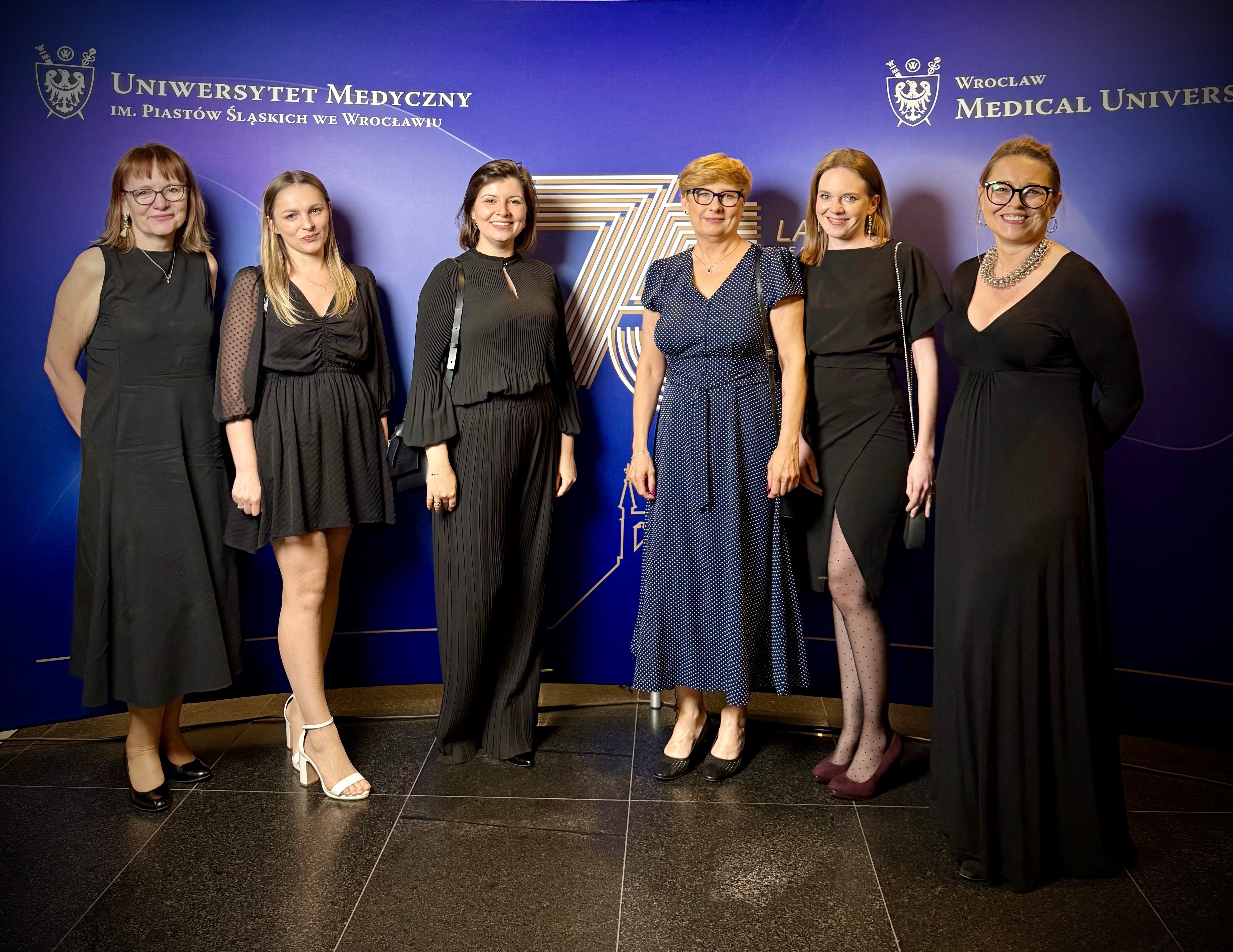 Zespół Katedry i Kliniki Geriatrii i Chorób Wewnętrznych na gali 75-lecia Uniwersytetu Medycznego we Wrocławiu. , na zdjęciu jest 6 lekarzy (kobiety) z od lewej: dr Małgorzata Szymala-Pędzik, lek. Monika Okołotowicz, lek. Katarzyna Kożuch - Sajdak, prof. dr hab. Małgorzata Sobieszczańska - Kierownik Katedry i Kliniki Geriatrii i Chorób Wewnętrznych, lek. Maria Rataj, mgr Marta Iskra. W tle logo 75 -lecia Uniwersytetu Medycznego we Werocławiu 